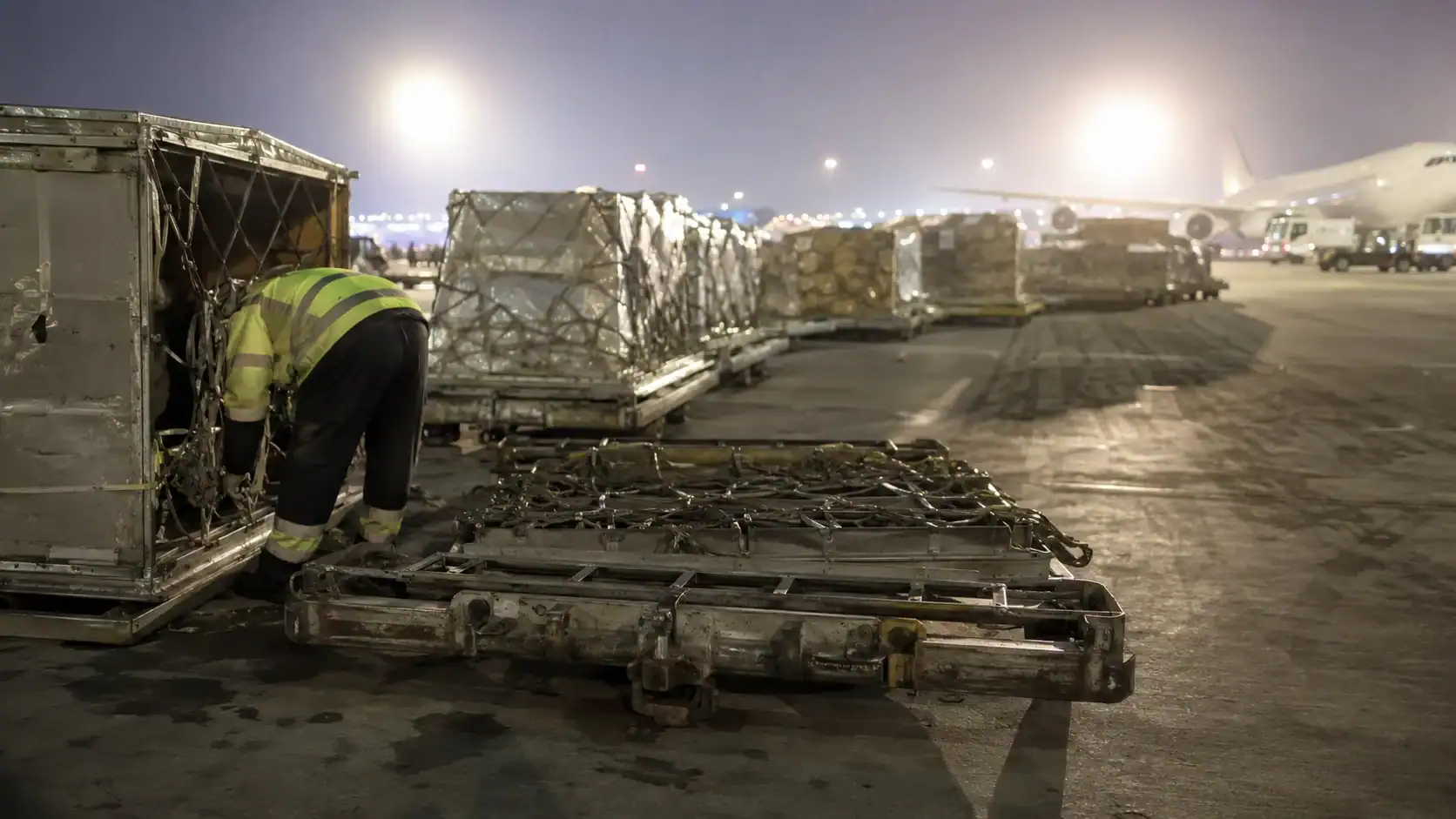 Ground crew worker securing cargo netting on an airport ramp at night, with freight pallets queued beside a wide-body cargo aircraft, illustrating the ground-handling stage where urgent shipments lose hours
