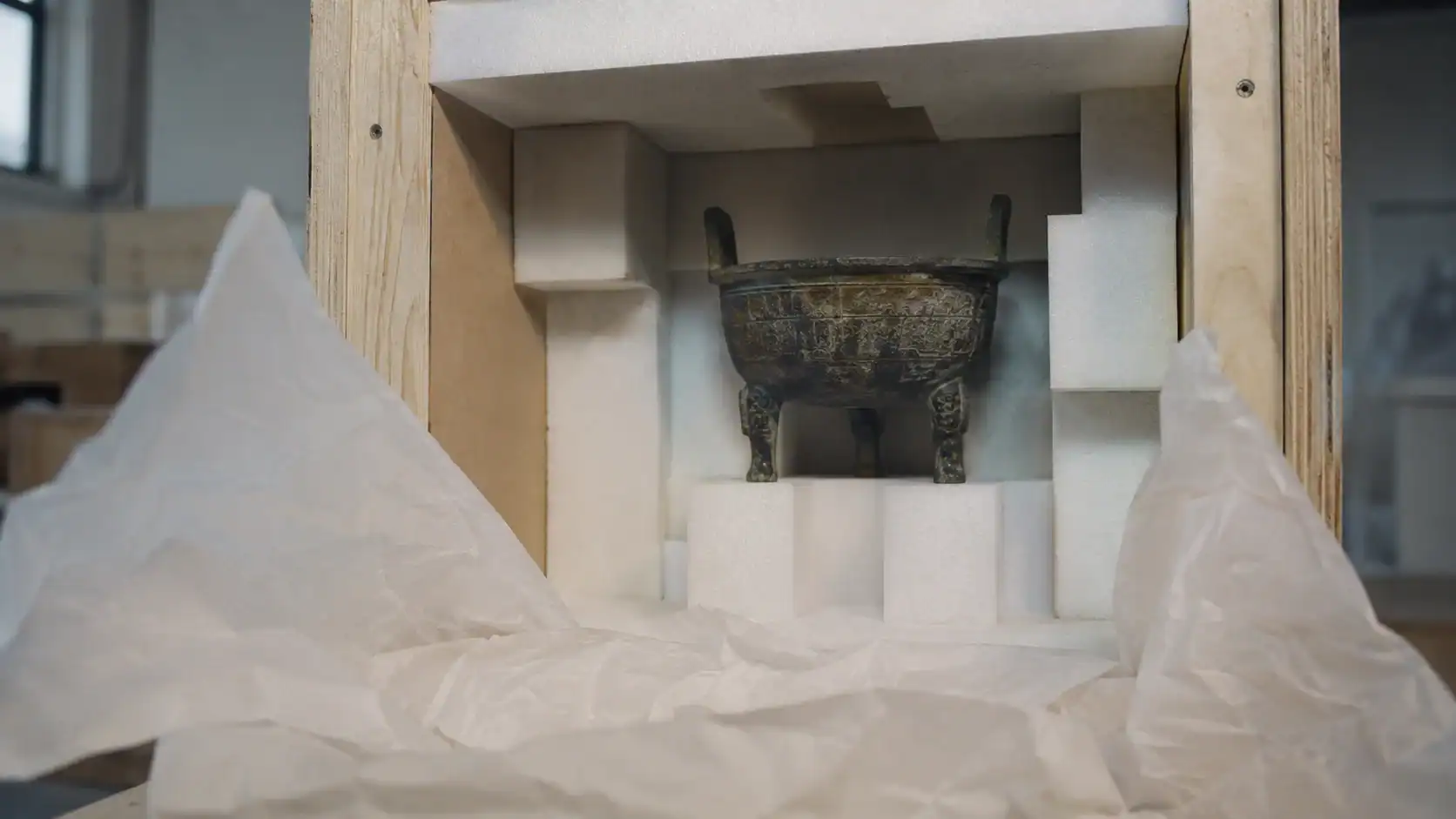 Antique bronze ritual vessel cushioned in cut foam blocking inside a wooden museum shipping crate with crumpled acid-free tissue paper, the kind of high-value fragile item that drives the case for specialist transit insurance