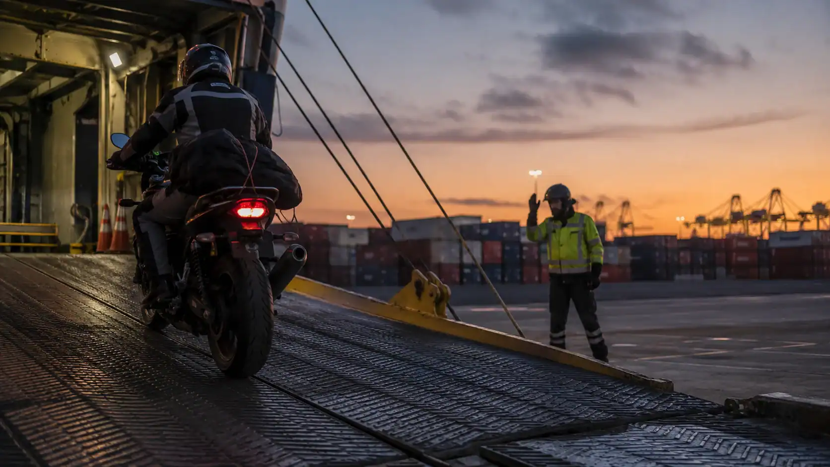 Motorcycle riding up a RoRo vessel loading ramp at an Australian port at dusk, guided by a hi-vis dock worker