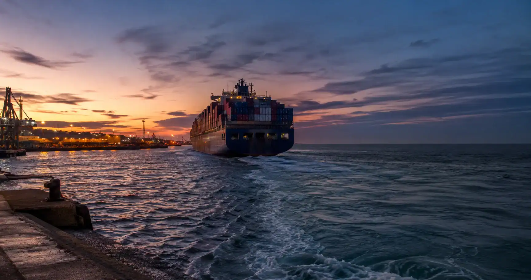 A loaded container ship departing a European port at dusk, gantry cranes silhouetted against the sunset sky, representing the long-haul sea freight route from France to Australia
