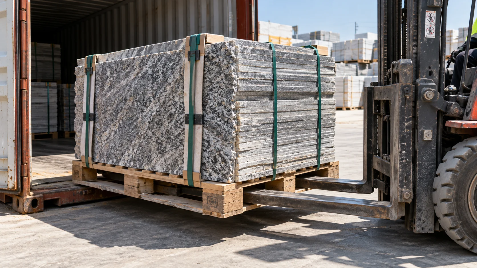 Stacked granite or porcelain tile slabs on a heavy-duty pallet beside an open container door, forklift tines just entering the frame to lift the pallet