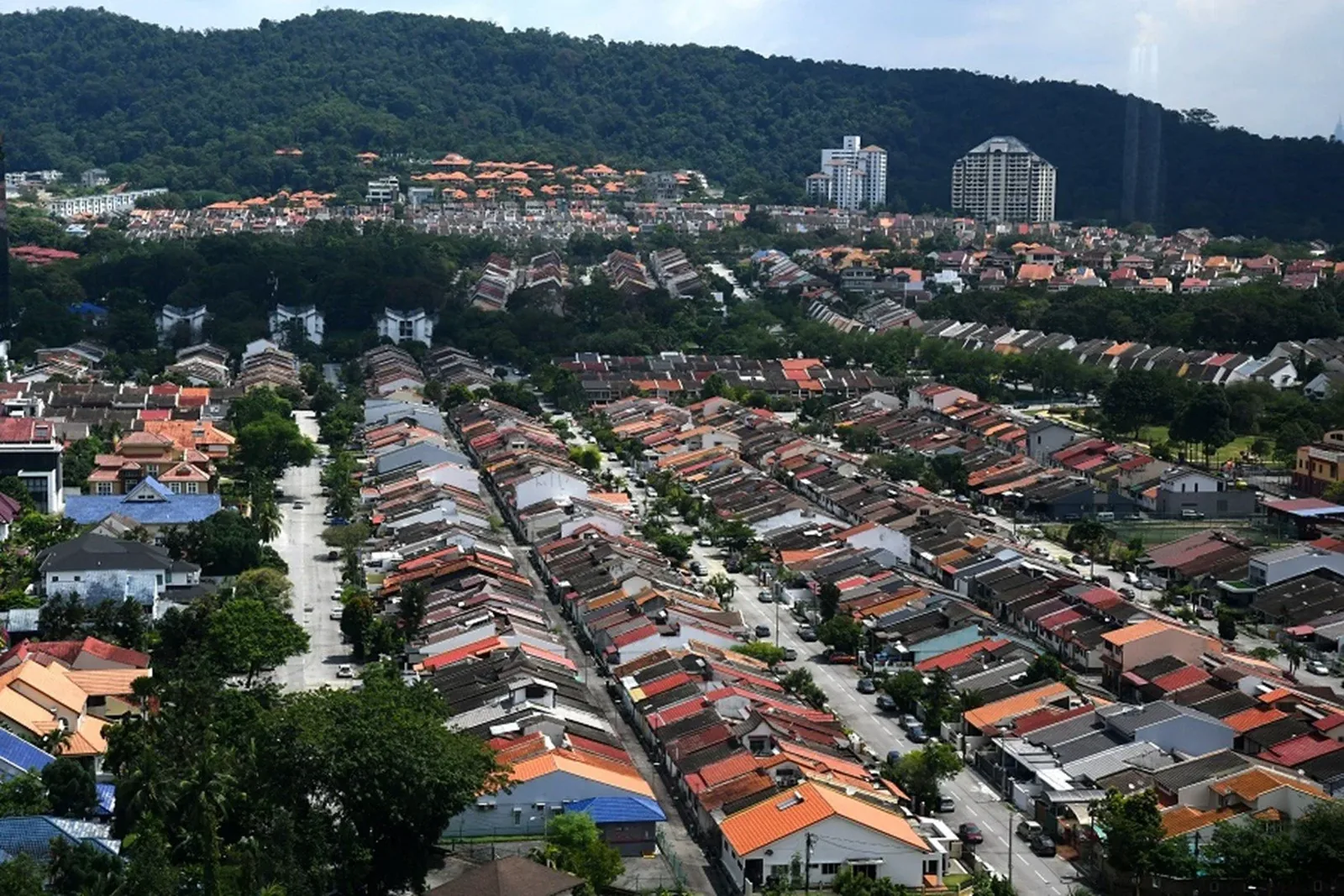 Aerial view of rows of terraced houses with tiled roofs below a forested hill and two apartment towers