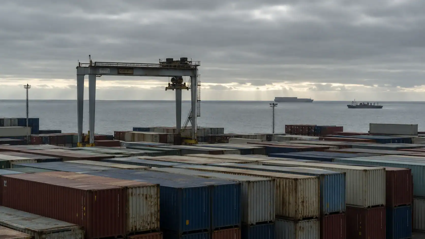 Elevated view of a densely packed container terminal yard with a gantry crane, cargo ships visible on the horizon under overcast skies