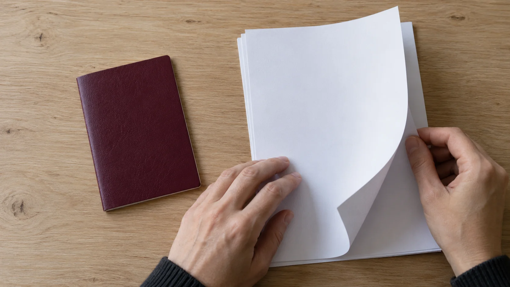 Hands turning a blank page from a paper stack beside a closed burgundy booklet, illustrating the paperwork steps of a transfer