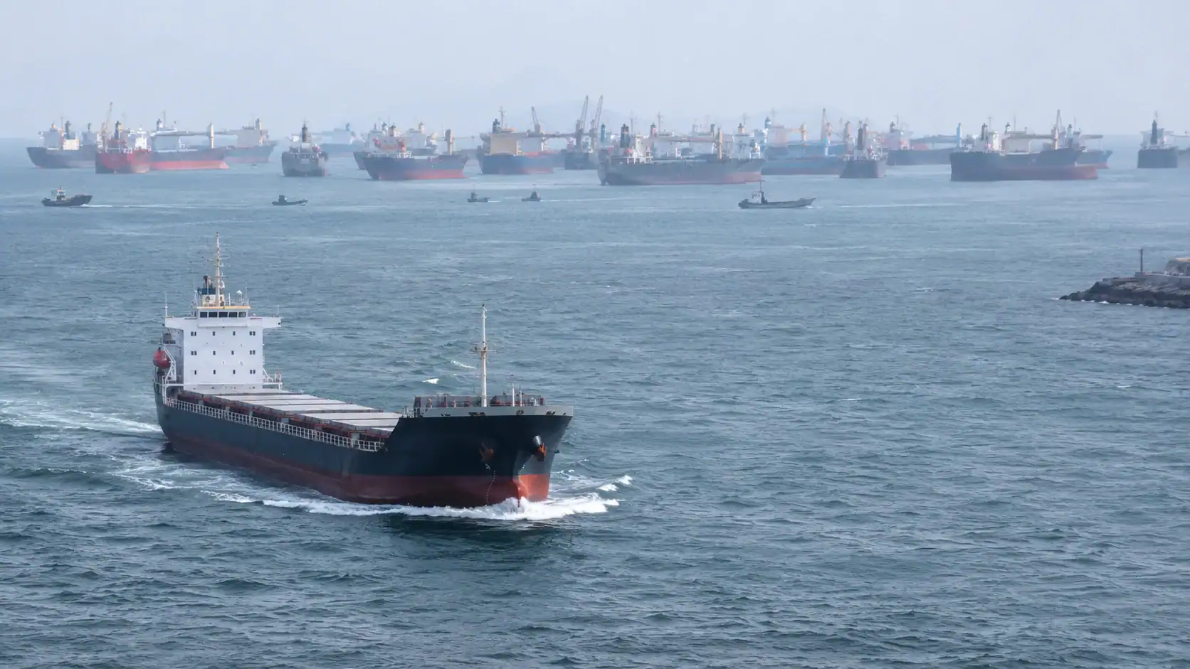 Bulk cargo ship underway past a dense queue of anchored vessels waiting off a congested port